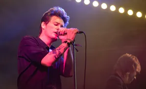 A male singer in a black shirt performs on stage, illuminated by stage lights and a string of lights overhead.