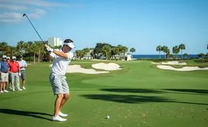 Golfer in white shirt and grey shorts swings club on a sunny day with spectators watching.