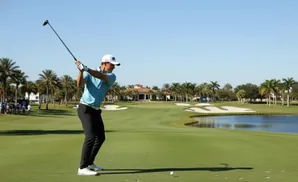 Golfer in mid-swing on a sunny day, with a golf course and palm trees in the background.