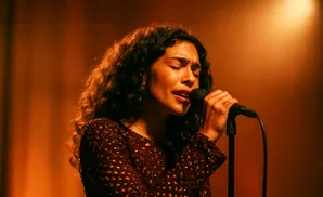 Woman with curly hair singing into a microphone on stage, bathed in warm, orange light.