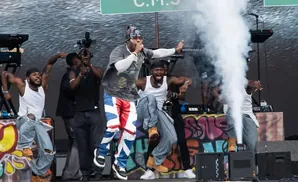 A male performer in colorful pants sings on stage with dancers and smoke effects.