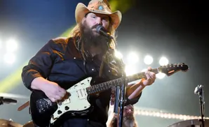A male singer in a cowboy hat plays a guitar on stage with bright lights behind him.