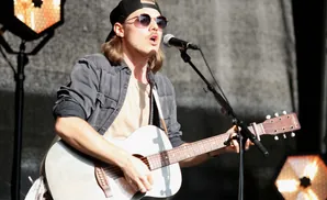 A male musician with sunglasses and a backward baseball cap plays an acoustic guitar and sings into a microphone on stage.