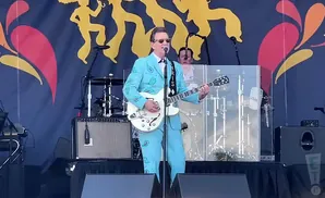 A man in a sparkly blue suit plays a white guitar on stage with a colorful backdrop.