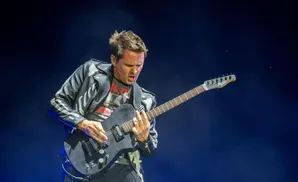 A male guitarist plays a black electric guitar on a dark blue stage.