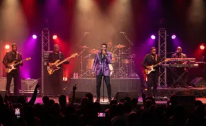 A band performs on stage under pink and blue lights, with a singer in a purple jacket center.