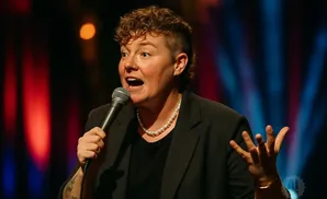 A comedian with curly hair and a pearl necklace holds a microphone while gesturing with their hands.