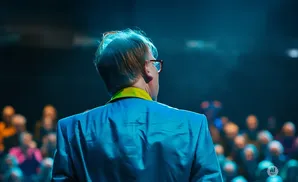 Man in a bright blue suit and yellow collar, facing away from the camera, with a blurred audience in the background.