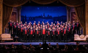 A choir of men in black suits and red vests perform on a stage with a starry night backdrop.