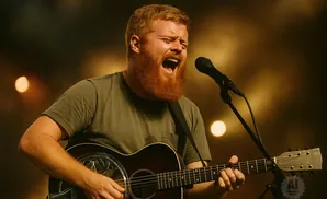 A red-bearded man sings into a microphone while playing an acoustic guitar on stage.