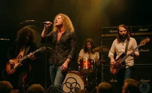 A band performs on stage: a singer with blonde curly hair, a guitarist, a drummer, and a bassist.