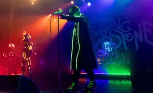 Musicians in futuristic neon costumes perform on stage with dramatic lighting and a band logo.