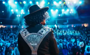 Man in a black hat and embellished jacket on stage facing a cheering audience.