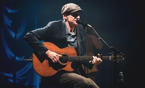 A man in a cap and suit plays an acoustic guitar and sings into a microphone on stage.