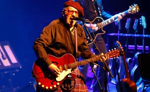 A musician in a red beret and tartan kilt plays a red guitar on stage with blue lighting.