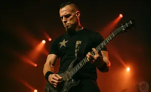 A male guitarist with a goatee and mohawk plays a black electric guitar on a dimly lit stage with orange spotlights.