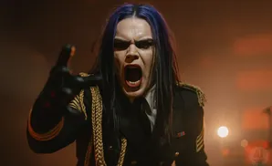 A person with blue hair and dark makeup screams while wearing a black military-style jacket with gold trim.