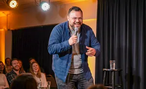 A comedian laughs into a microphone on stage while a crowd watches.