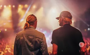 Two men facing away from the camera, looking at a blurred concert crowd and stage lights.