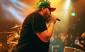 A man with a spiked hat sings into a microphone on stage with a drummer in the background.