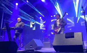 Two musicians performing on a brightly lit blue stage. One plays a keyboard, the other a guitar.