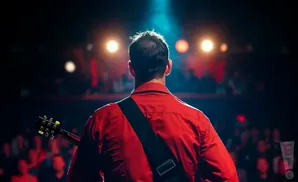 A man in a red shirt with a guitar strap stands on a stage, facing away from the camera, with bright lights and an audience behind him.