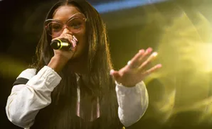 A woman with long dark hair and pink cat-eye glasses sings into a microphone, her hand raised.