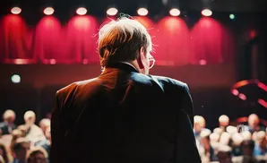 Man in a suit facing an audience, with red stage lights behind him.
