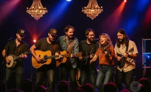 A band of six musicians performs on stage, playing guitars, a banjo, a mandolin, and a fiddle under dramatic lighting.
