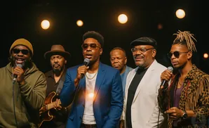 A Black male band performs on stage with microphones and instruments, lit by warm spotlights.