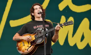 A man with long brown hair plays a sunburst hollow-body guitar and sings into a microphone on a stage.