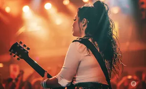 A woman with her back to the camera plays an electric guitar on a brightly lit stage.