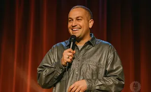A man with a shaved head, wearing a shiny silver shirt, smiles while holding a microphone on stage.