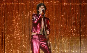 A singer in a sparkly red outfit performs on stage against a shimmering gold fringe curtain.