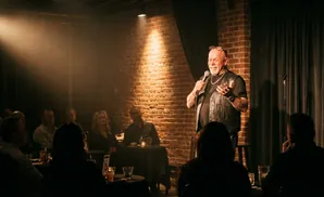 A man tells jokes on stage to a dark room full of people.