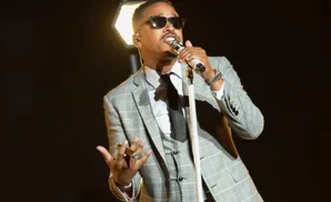 Man in a plaid suit and sunglasses sings into a microphone.