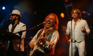 Three men perform on stage, one playing guitar and singing, another at a keyboard, and the third singing into a microphone.