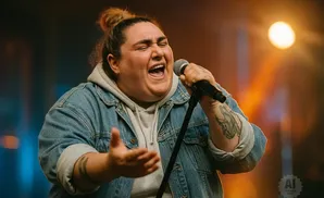 A singer with a bun hairstyle, wearing a denim jacket, belts into a microphone on a dimly lit stage.