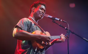 A Black man smiles while playing a ukulele and singing into a microphone on a stage with colorful lighting.