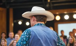 A man in a white cowboy hat and denim vest faces away from the camera, with a crowd blurred in the background.