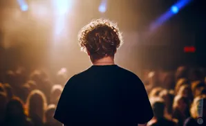 A person with curly hair stands on a stage facing a blurry crowd, illuminated by warm stage lights.
