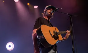 Musician in a cap sings into a microphone while playing an acoustic guitar.