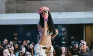 A female performer in a pink hat sings into a pink microphone in front of a blurred crowd.