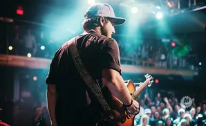Guitarist with a cap plays on stage with bright lights and a cheering crowd.