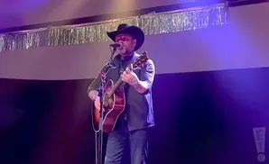 A man in a cowboy hat plays an acoustic guitar on stage, illuminated by purple lights.