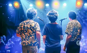 Three men on stage with microphones, backlit by blue and yellow lights, facing an audience.