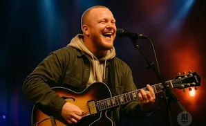 A bald man with a beard laughs while playing a guitar and singing into a microphone on stage.