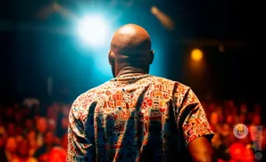 A bald man in a colorful patterned shirt stands on stage, facing away from the camera, under a bright spotlight.