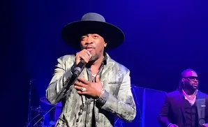 A man in a hat and patterned suit sings into a microphone on stage with a blue background.