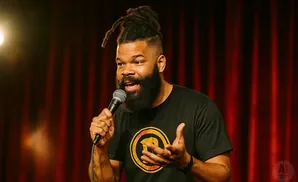 A man with dreadlocks and a beard holds a microphone and speaks into it while gesturing with his hands.